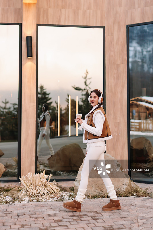 一位漂亮的女孩穿着保暖的衣服，边走边喝着咖啡，手中还拿着一杯热饮，享受着户外的自然风光。图片素材