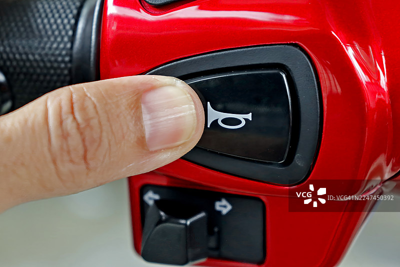 特写：手指按下黑色喇叭按钮，该按钮带有风格化的喇叭图标，位于摩托车车把明亮的红色外壳上。图片素材