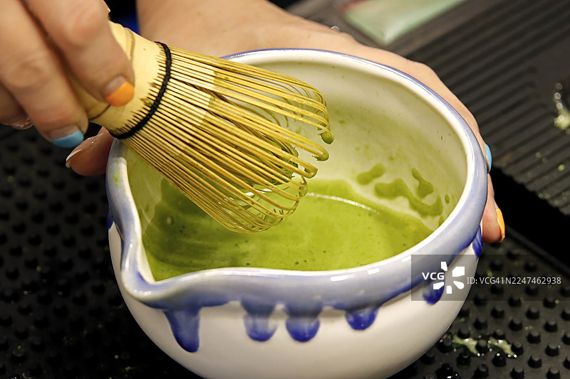特写：一只手正用传统的竹制茶筅在一个手绘的日式陶瓷碗里搅打翠绿的抹茶图片素材