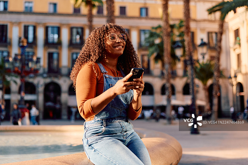 一位年轻女子在巴塞罗那城市广场上微笑着使用智能手机图片素材
