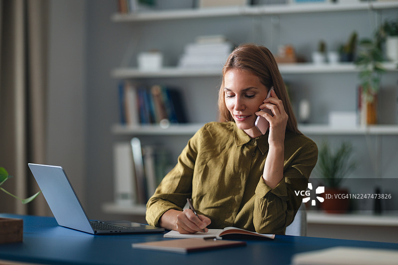 女商人在家里的办公室办公，正在打电话、写笔记，旁边有笔记本电脑和绿植。图片素材
