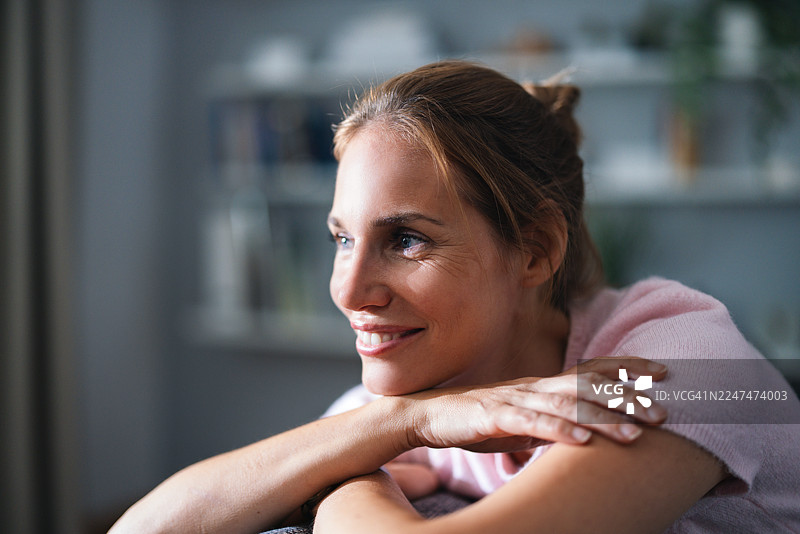 穿着粉色舒适毛衣的微笑女性，在家中办公室将下巴搭在叠起的胳膊上图片素材