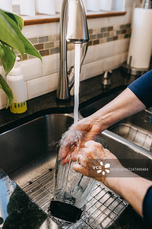 一个人正在厨房的水槽里，在流水下冲洗搅拌机容器。图片素材