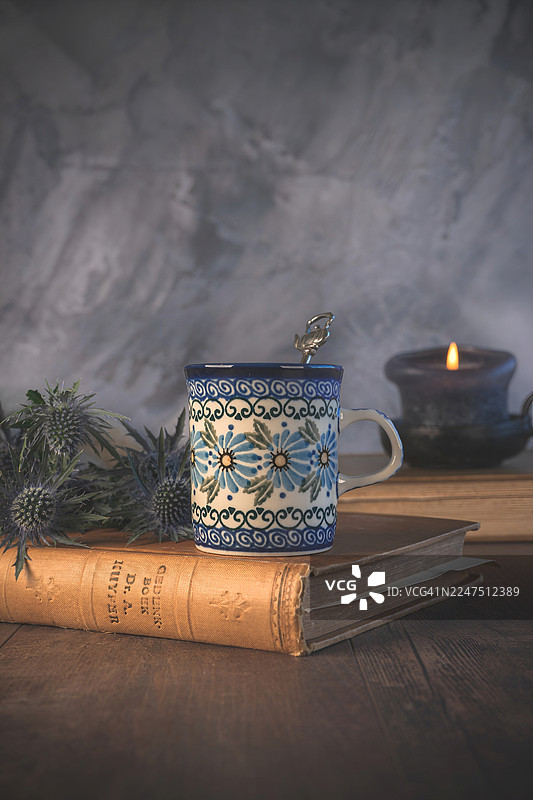 舒适的静物，包含一本古董书、一个花纹咖啡杯、一支点燃的蜡烛和蓟花，摆放在质朴的木桌上图片素材