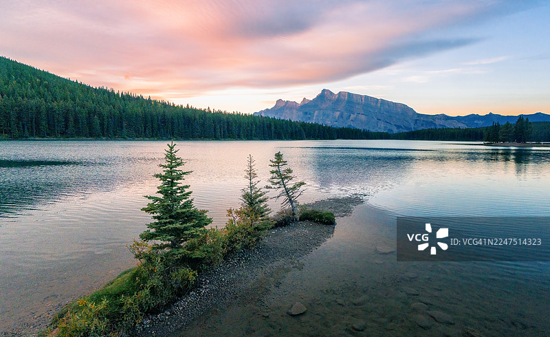 加拿大卡尔加里班夫国家公园，夕阳下的宁静湖泊倒映着远山和绚烂天空，前景是伸入湖中的小片松树林。图片素材