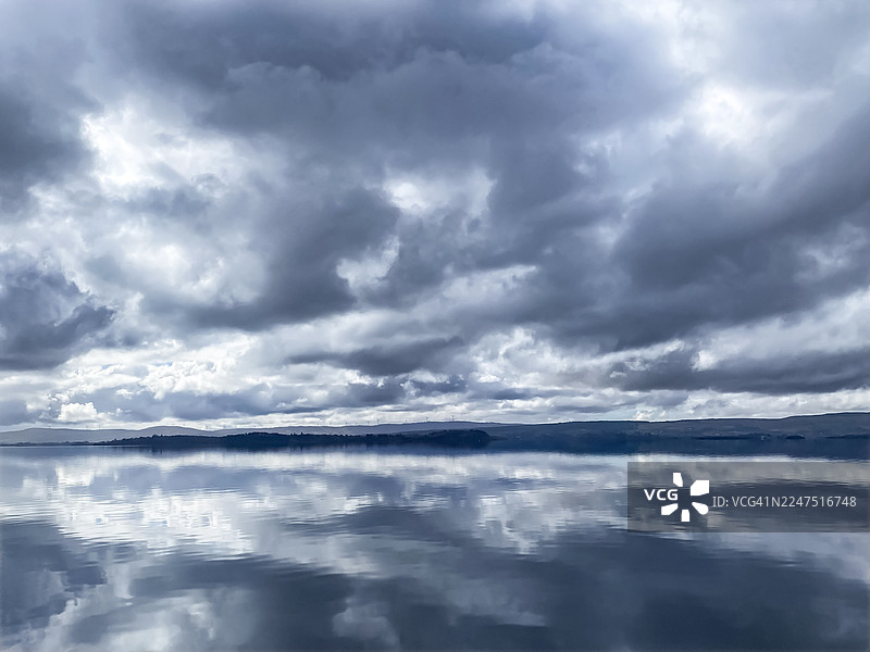 阴天之下，平静的湖水倒映着戏剧性的多云天空和远处的山丘图片素材