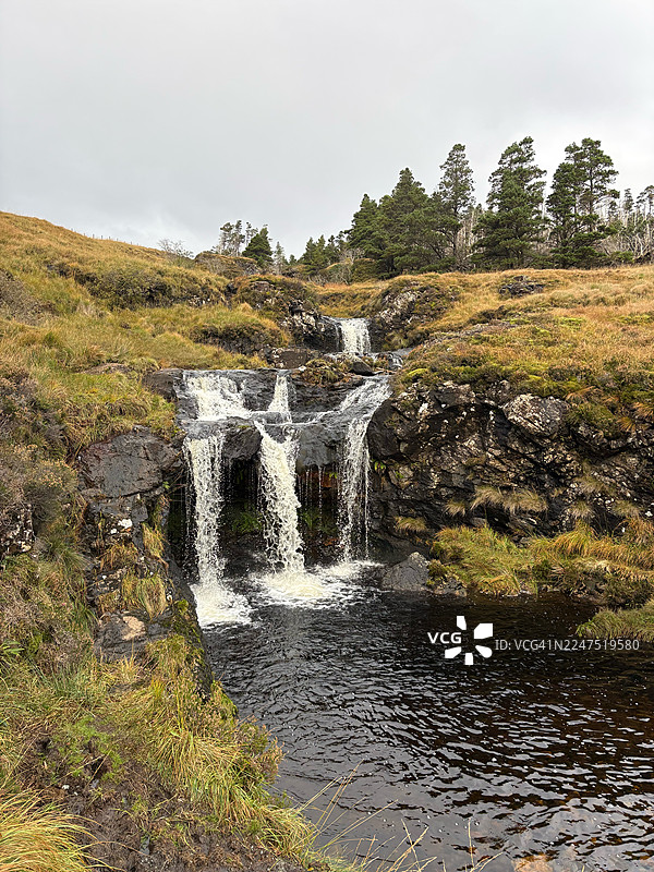 英国史密斯顿，瀑布沿着布满岩石的地形倾泻而下，上方是多云的天空，周围环绕着郁郁葱葱的绿色和金色的草地，仙境般的景色图片素材