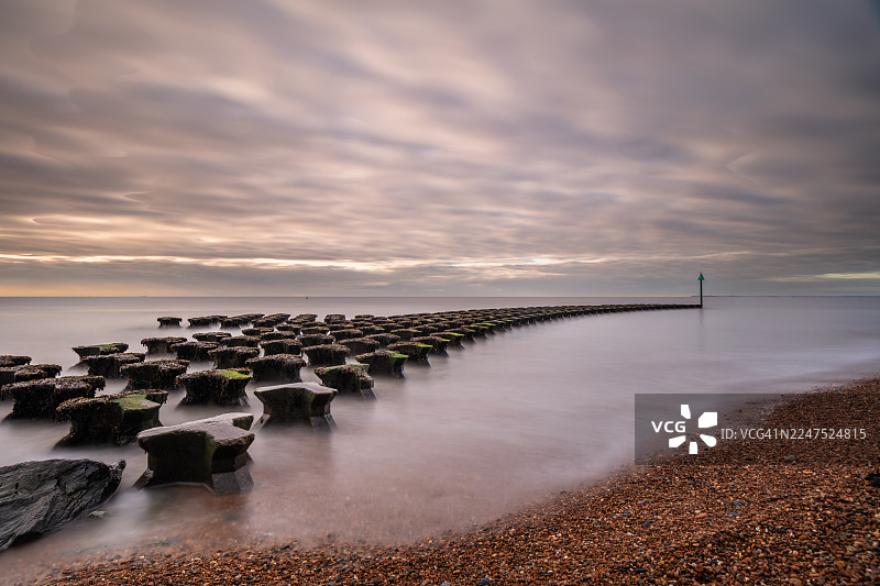英国东萨福克郡费利克斯托，夕阳下的宁静海滩与混凝土防波堤的长曝光景象图片素材