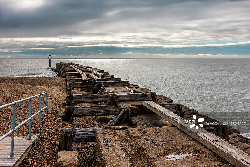 多云天空下，一道饱经风霜的木制丁坝平静地伸入海中，构成一幅风景画。图片素材