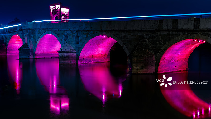 夜幕下的历史悠久桥梁，河流之上，霓虹粉色灯光与车流光轨交织图片素材