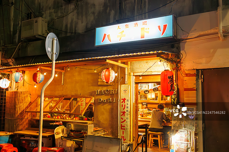 日式居酒屋外观。设有露天吧台，展示着老式踏板车。游客正进入店内，偶尔有出租车经过。图片素材