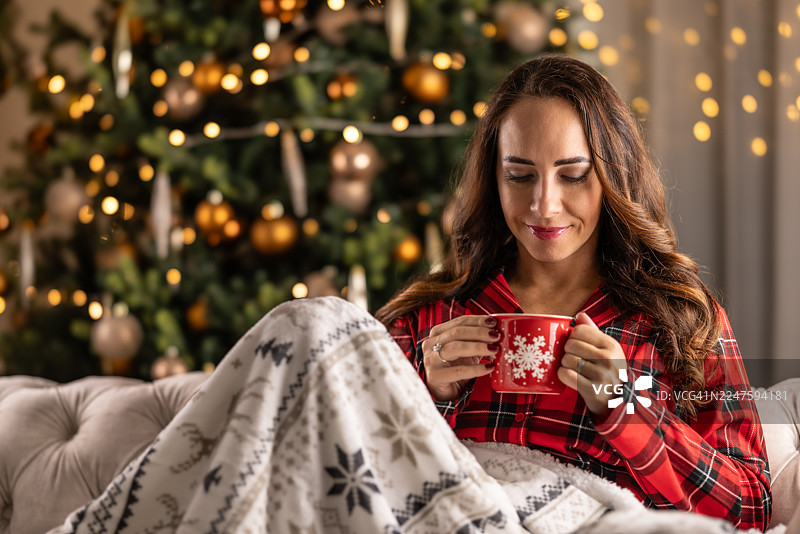 一位可爱的棕发女子穿着红色格子睡衣，裹着带有圣诞图案的温暖毯子。她手中拿着一个装有热乎乎的圣诞潘趣酒的红色马克杯，享受着酒的香气。图片素材