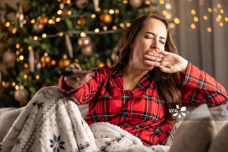 圣诞节的宁静时光，盖着毯子看电视的努力直到深夜，女子因疲倦而打哈欠，用手捂着嘴，背景中圣诞树闪耀。图片素材