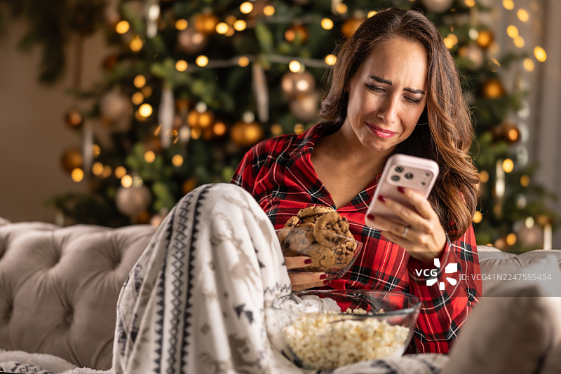 家中的圣诞气氛无法让她开心起来，身穿睡衣的女子坐在闪闪发光的圣诞树前，眼中含泪，手中拿着手机，试图用一碗爆米花来抚慰悲伤。图片素材