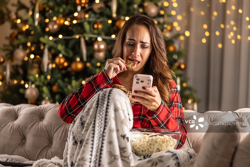 家中的圣诞气氛无法让她开心起来，身穿睡衣的女子坐在闪闪发光的圣诞树前，眼中含泪，手里拿着手机，并试图用一碗爆米花来缓解悲伤。图片素材