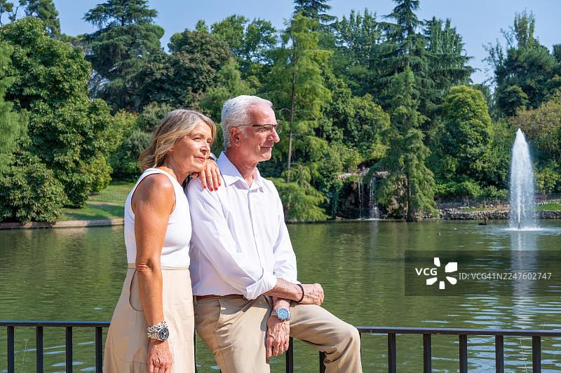 马德里，西班牙，欧洲。一对年迈夫妇站在宁静的湖边，背景是美丽的绿色公园和喷泉，在 retiro 公园享受浪漫约会。图片素材