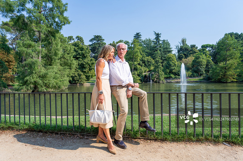 夏日晴朗的一天，马德里，西班牙，欧洲，丽池公园内，一对年长夫妇依偎在栅栏旁，在闪闪发光的湖泊、喷泉和绿色树木的映衬下，享受着悠闲浪漫的约会。图片素材
