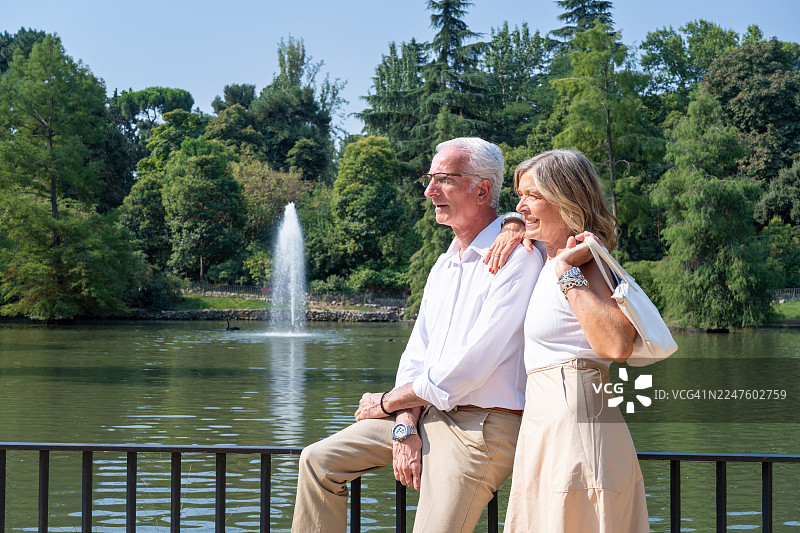 马德里丽池公园水晶宫湖畔,一对成熟的夫妇正在拥抱,享受着阳光明媚的一天,欣赏着湖光景色,展现了浪漫与悠闲的退休旅行生活。图片素材
