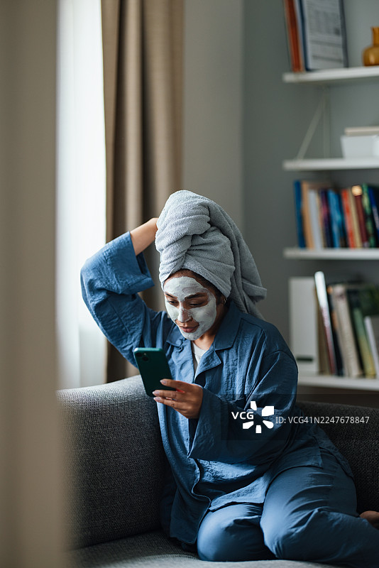 身着蓝色浴袍的放松女性在家中沙发上浏览智能手机，脸上敷着面膜图片素材