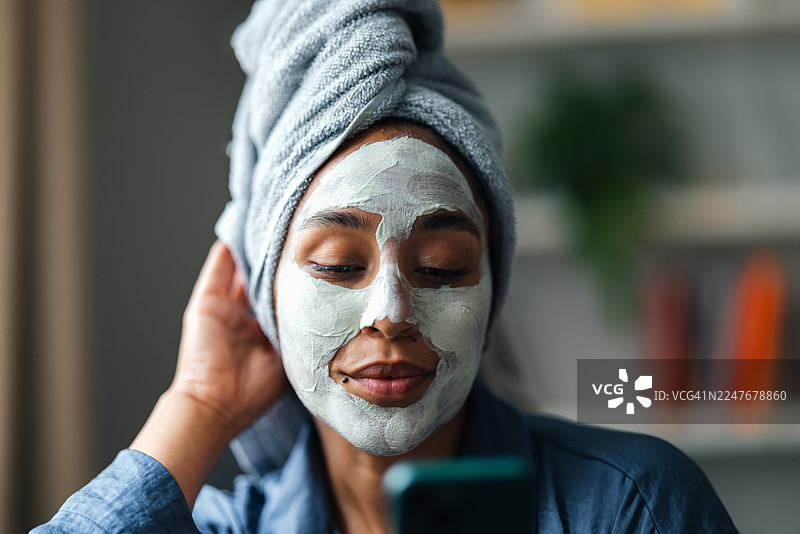 女子在家敷着面膜、裹着浴巾，一边看手机一边进行自我护理图片素材