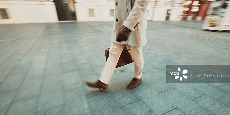 一个穿着米色外套、提着棕色公文包的成年男子走在城市的人行道上图片素材