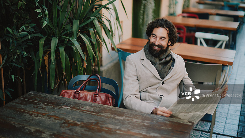 英俊的成年男子在户外咖啡馆阅读报纸，周围有植物和节日装饰。图片素材