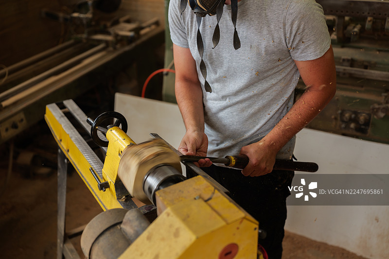 木匠戴着防护口罩，在布满灰尘的旧工坊里，使用传统手工工具在车床上加工木材，塑造木件。图片素材