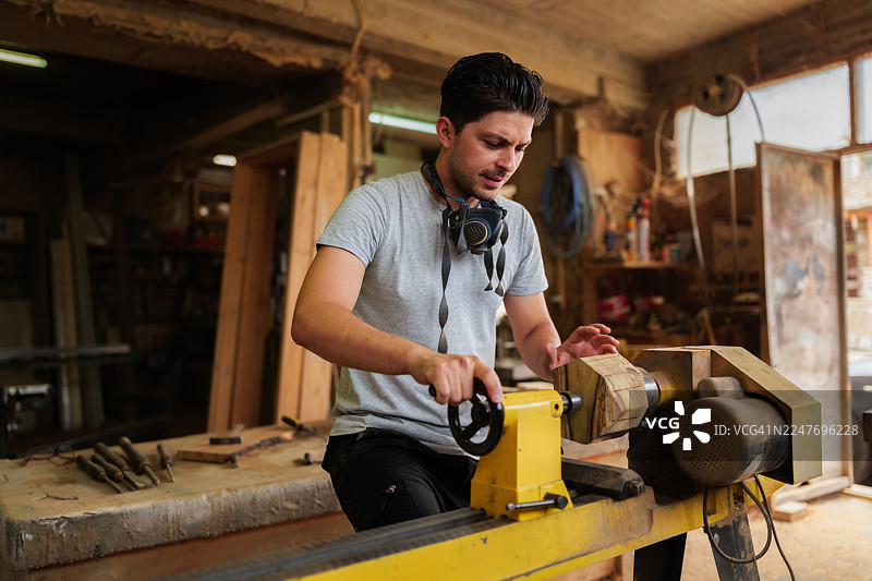 一位戴着防尘口罩的年轻木匠，在充满工具和木屑的旧工坊里，熟练地操作车床打磨木材。图片素材
