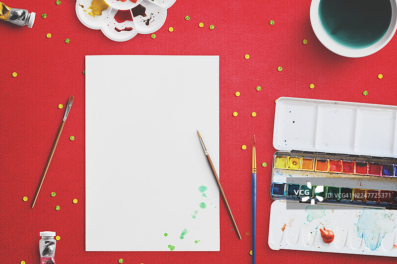 充满活力的俯视构图，展示了摆放在撒有黄色和绿色亮片的鲜艳红色背景上的各种艺术用品。图片素材