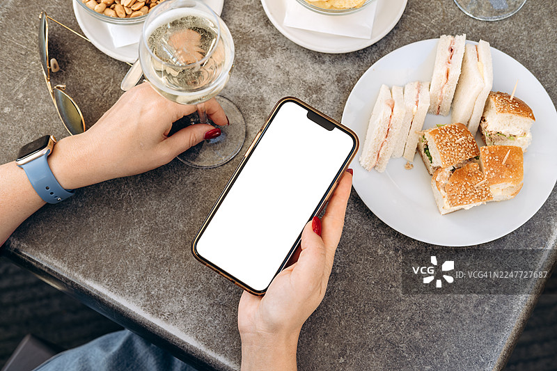 咖啡馆场景中，女子手持带有空白屏幕的智能手机模型，周围环绕着精致的饮品和美味的开胃菜，非常适合设计使用图片素材