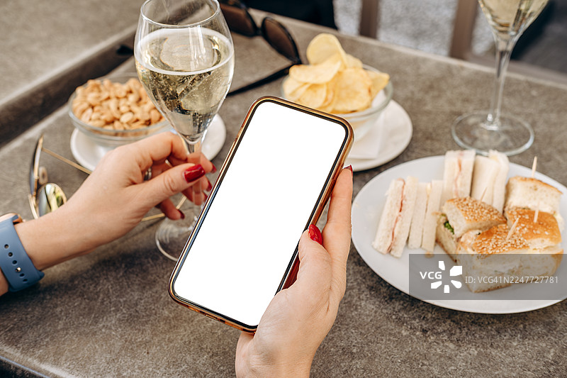 咖啡馆场景中，女子手持带有空白屏幕的智能手机模型，周围是精致的饮品和美味的开胃菜，非常适合设计用途图片素材