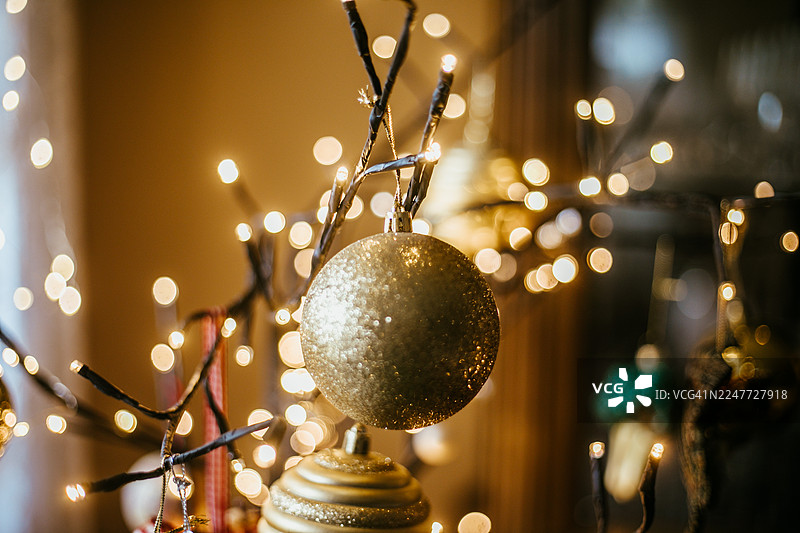 图片捕捉到迷你串灯与树枝交织的温暖氛围光芒。一个闪闪发光的金色大装饰球醒目地悬挂在前景中，其下方可见另一个有肋条纹的金色装饰球。柔焦效果营造出一种魔幻图片素材