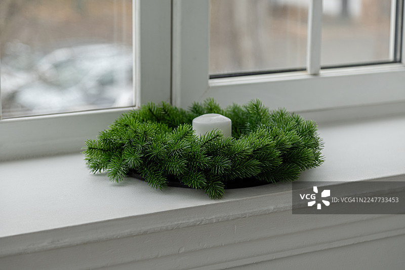 一个白色窗台上放着一个小巧的人工绿色松树花环，上面点缀着一支蜡烛。图片素材
