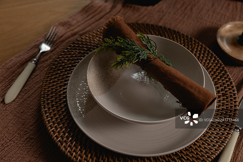 一个卷起的棕色餐巾，上面放着一小簇绿色人造嫩枝，放在一个陶瓷碗里，碗放在一个盘子上，盘子放在一个圆形的餐垫上，餐垫介于一把叉子和一把刀之间。图片素材
