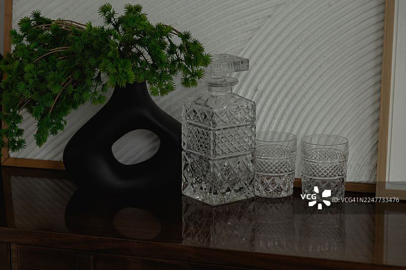 一个黑色花瓶，里面插着绿色植物，一个方形切割的醒酒器和两个酒杯摆放在棕色桌子上。图片素材