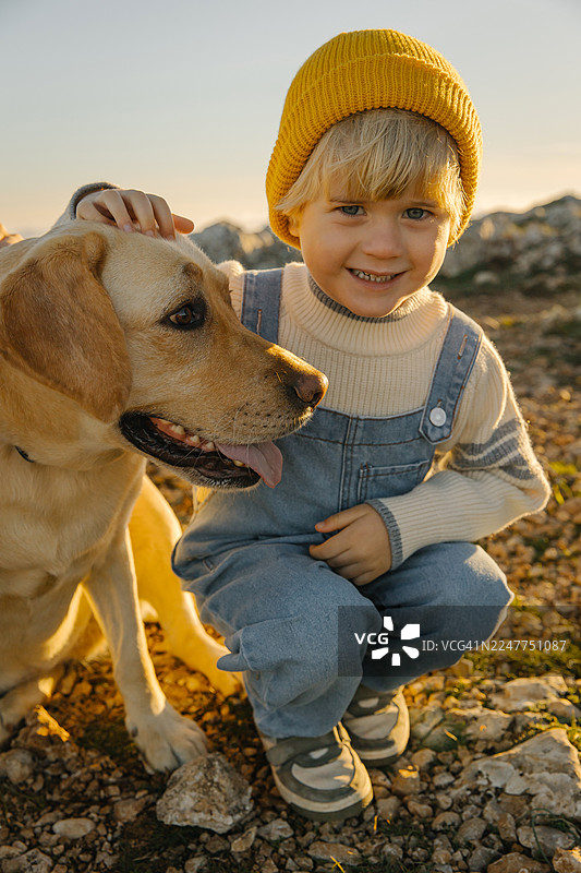 阳光明媚的户外，一个戴着黄色绒帽的快乐孩子和一只狗图片素材