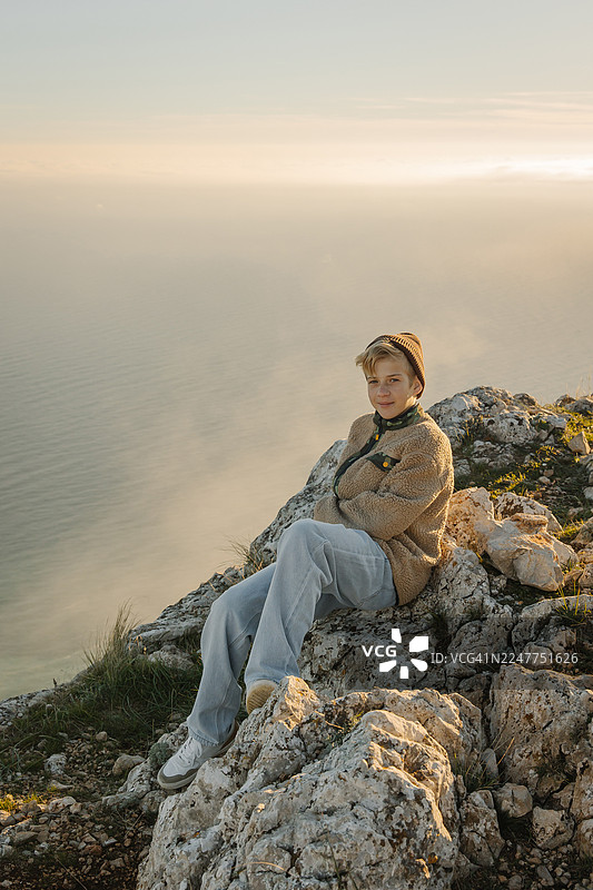 日落时分，一位沉思的女子坐在悬崖边，眺望着平静的大海。图片素材