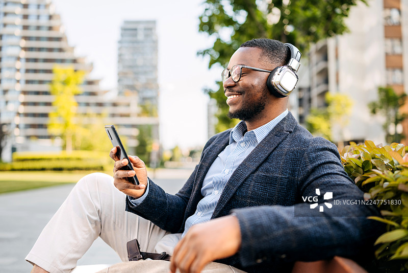 一位面带微笑的商人正坐在城市市中心的户外长椅上使用手机。图片素材
