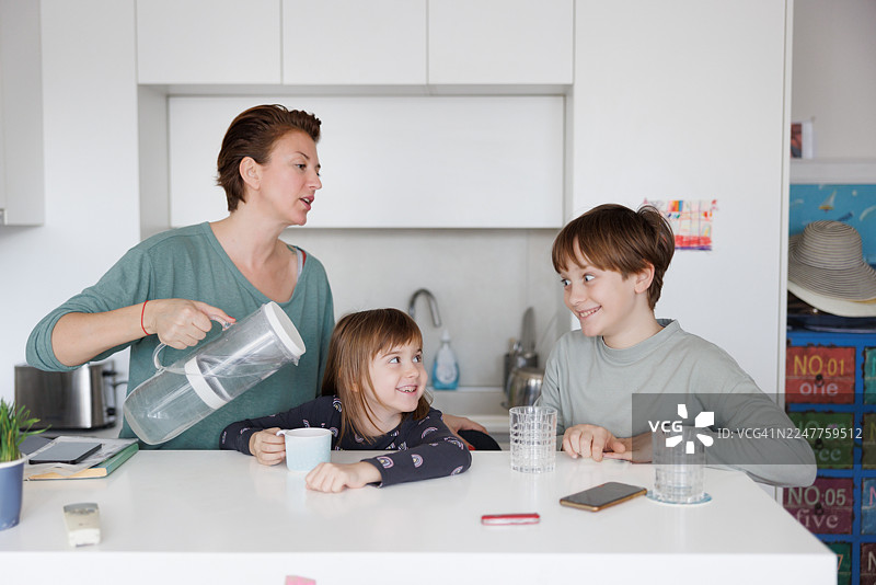 中年女子在厨房柜台旁，抱着水罐，和她的小女儿及儿子站在一起图片素材