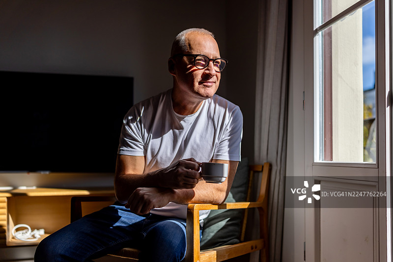 阳光明媚的客厅里，一位年长男士正坐在窗边悠闲地喝着咖啡图片素材