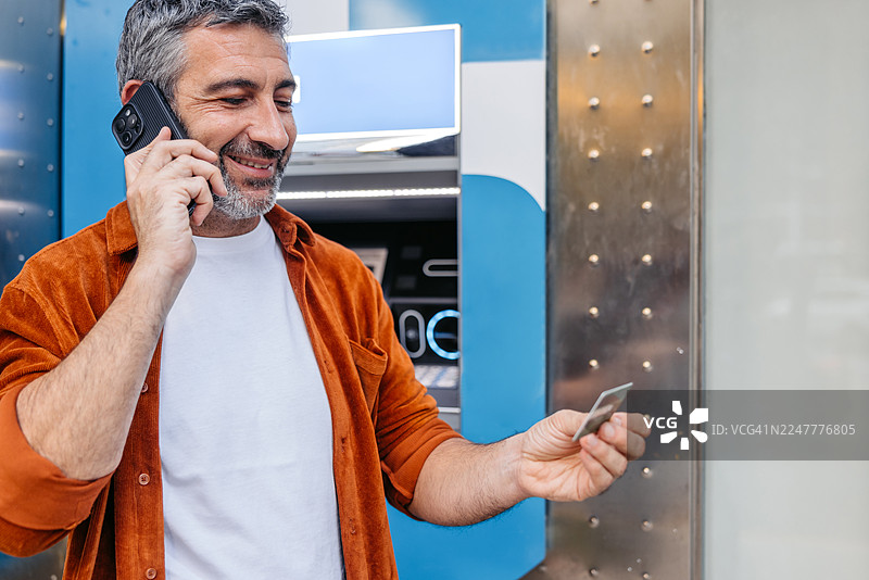 西班牙巴塞罗那，一位成熟的男士在自动取款机前边打电话边拿着信用卡。图片素材