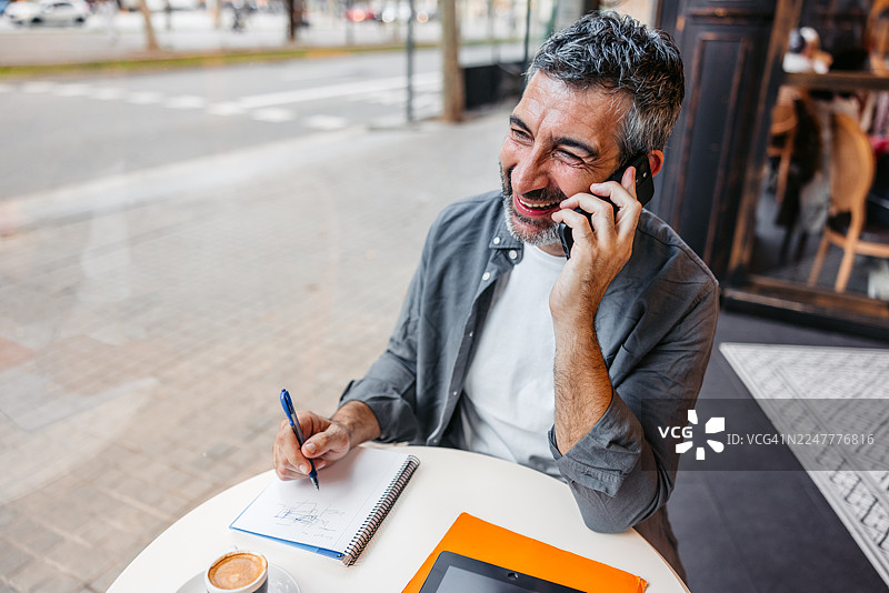 在巴塞罗那的一家咖啡馆里，一位成熟的男士一边喝着咖啡，一边在笔记本上写字并讲着电话。图片素材