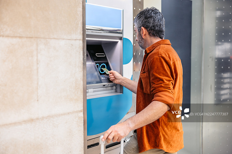 一位成熟的男士在西班牙巴塞罗那的自动取款机上，携带带轮行李箱，使用信用卡取款。图片素材