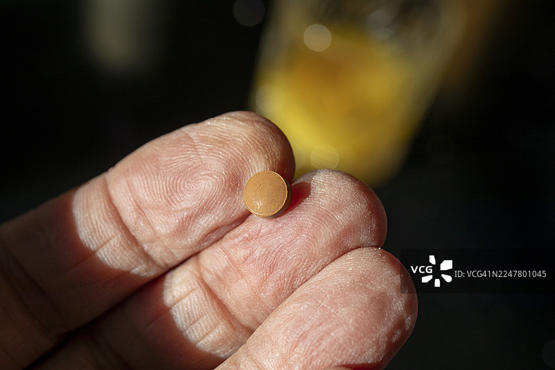 服用铁补充剂——一颗富马酸亚铁药片放在一位老年男性的手指上，背景是橙汁杯图片素材