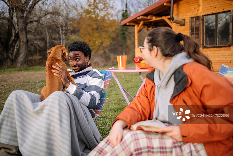 秋日暖阳下，人们坐在小屋前，悠闲地与宠物玩耍。图片素材