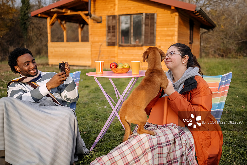 秋日暖阳下，人们在小屋前放松，与宠物玩耍并用手机拍照图片素材