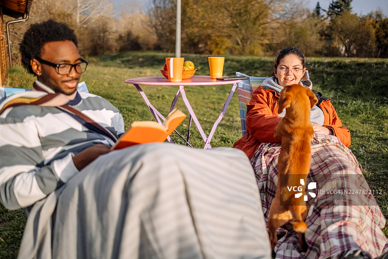 秋日暖阳下，人们在小屋前悠闲地放松，与宠物玩耍，同时阅读书籍。图片素材