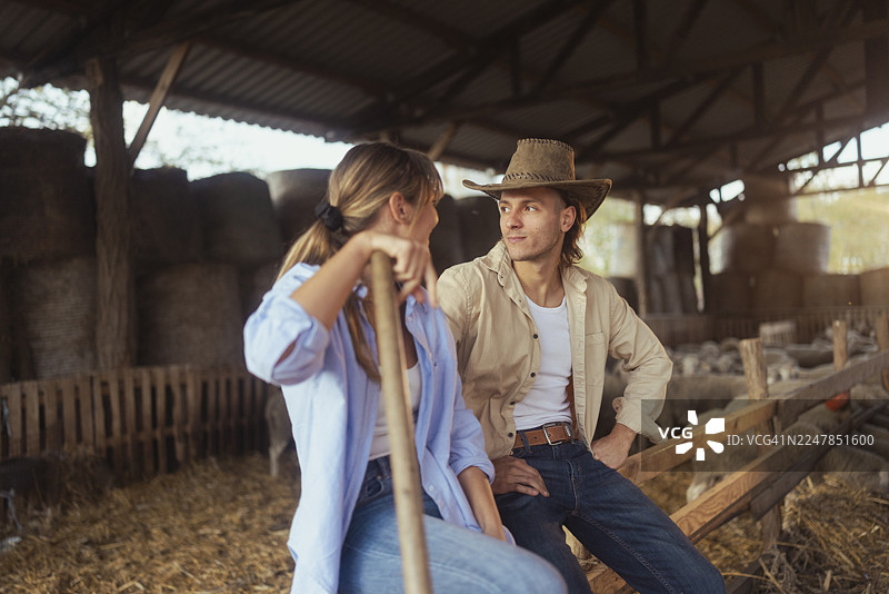 两名农夫，一男一女，穿着休闲农场服装，在羊圈里坐着并微笑着，背景中有干草和羊，自然光从窗户透入。图片素材