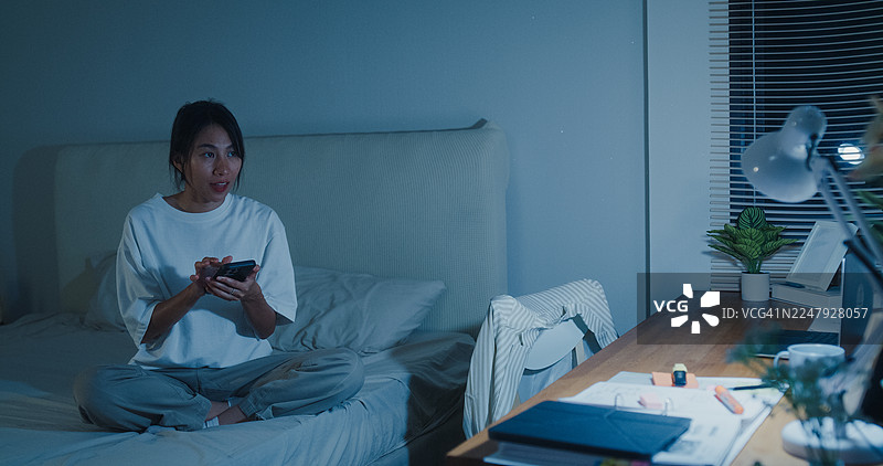 年轻的亚洲女性在家中卧室的夜晚，使用智能手机开关灯，坐在床上。智能家居技术在日常生活中的应用概念。图片素材