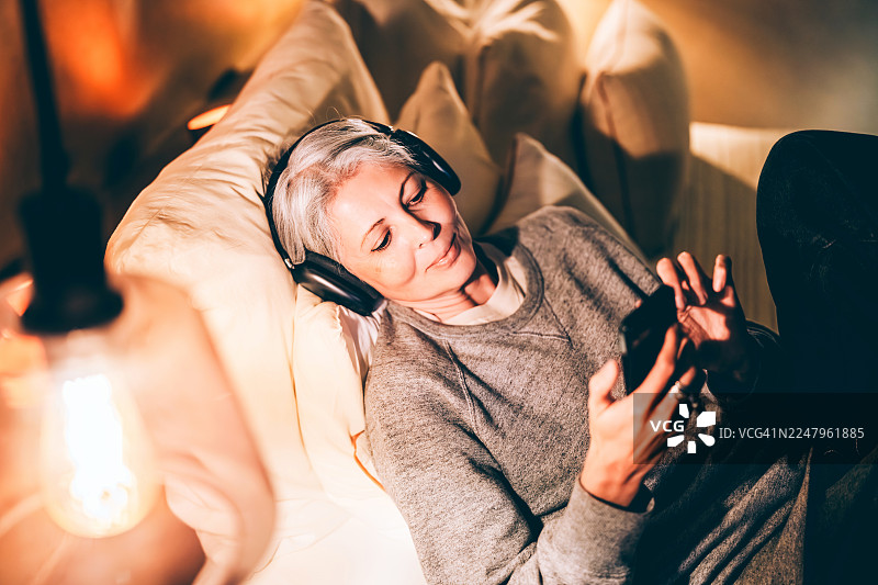 居家放松时刻：女子戴着耳机，一边听音乐一边使用智能手机，舒适地躺在柔软的床上，在温馨舒适的客厅环境中享受休闲的个人时光。图片素材